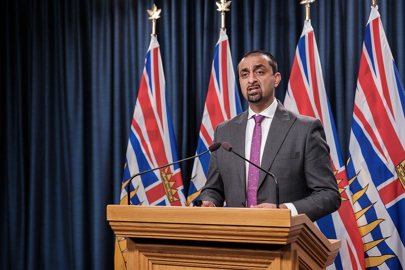 Photo of Housing Minister Ravi Kahlon standing at a podium in front of four BC flags