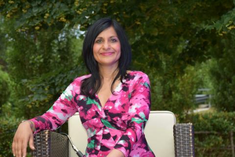 A person with long black hair, dressed in a pink patterned dress, sitting in a chair and smiling into the camera