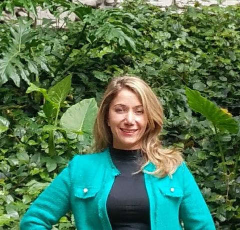 Yenny Vega Cardenas, a blonde woman dressed in a black mock neck shirt and a bright blue jacket, is photographed in front of a wall of greenery and smiles into the camera
