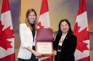 Two women stand in front of three Canadian flags holding a certificate between them